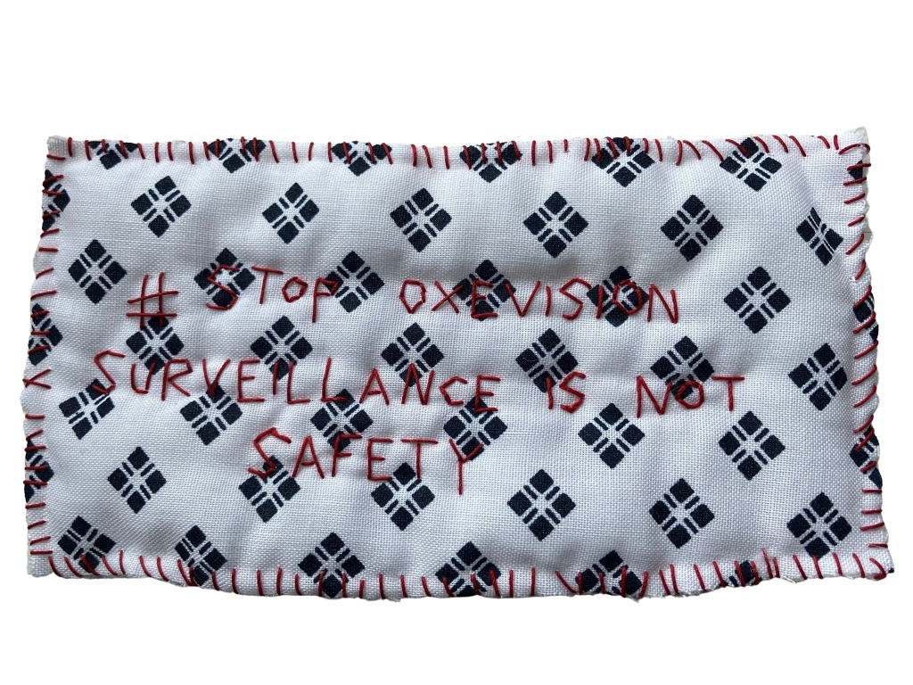 Image of a stitched piece of hoptial gown fabric with the words stitched in red thread reading "# stop Oxevision // surveillance is not safety" there are red stitches around the edge of the piece of fabric